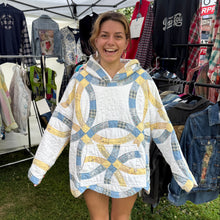 Load image into Gallery viewer, A woman smiling outdoors under a white market canopy, wearing a loose white quilted hoodie with large abstract blue and yellow geometric patterns. Clothing racks with various garments are visible behind her on grass.
