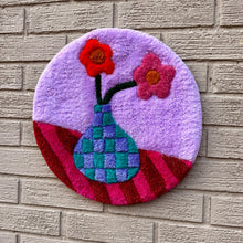 Load image into Gallery viewer, A circular tufted wall hanging featuring a purple background, a blue-green checkered vase, and two flowers in red and pink, displayed against a light gray brick wall.
