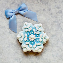 Load image into Gallery viewer, A snowflake-shaped sugar cookie decorated with light blue and white icing, placed on a gray marble surface next to a light blue satin bow.
