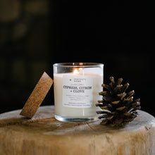 Load image into Gallery viewer, A lit candle in a clear glass jar sits on a rustic wooden stump, with a cork stopper beside it and a pinecone in front. The label reads 'CYPRESS, CITRON + CLOVE'.
