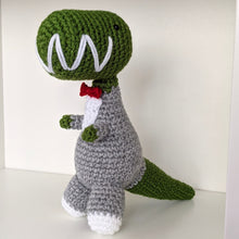 Load image into Gallery viewer, A handmade crocheted dinosaur toy wearing a gray tuxedo with a red bow tie, white shirt details, and a green head and tail, standing on a white surface against a plain light background.
